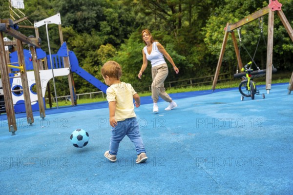 Happy toddler playing soccer with his mother at the playground on a sunny summer day, enjoying