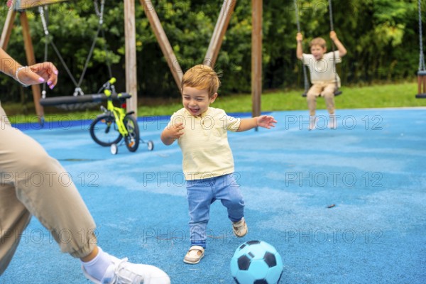 Happy toddler boy kicking soccer ball at playground with family, enjoying a playful moment on a