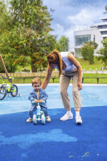 Young mother guiding her son as he learns to ride a balance bike on a playground featuring a soft