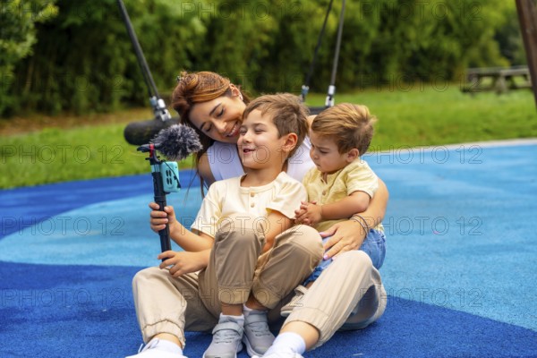 Mother and two sons are sitting on playground's rubber surface, holding a smartphone attached to a