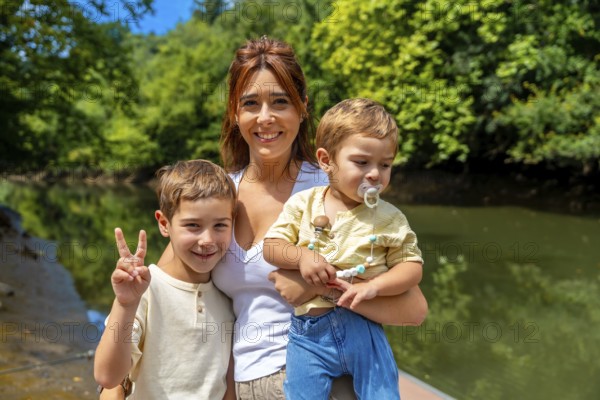 Caucasian mother joyfully holding her two sons near a river on a sunny summer day, with one son