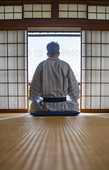 Young man wearing kimono sitting in traditional Japanese living room with tatami mats and shoji