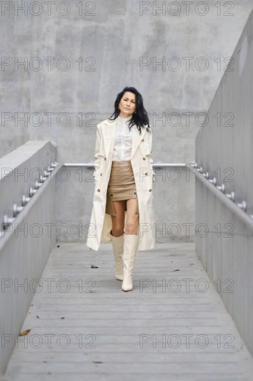 A woman strolls down a sleek walkway, wearing a beige trench coat over a white blouse and a stylish