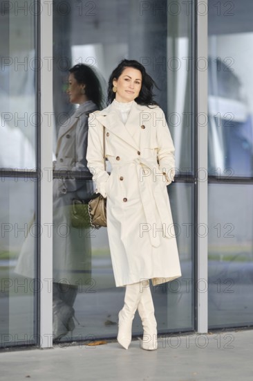 A woman showcases her autumn street style, wearing a cream coat and boots while leaning against a