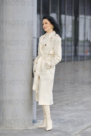 A confident woman stands next to a concrete pillar, dressed in a cream trench coat and matching