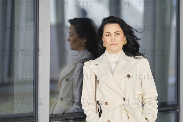 A woman stands confidently in a white trench coat, with her hair elegantly flowing on the wind. She
