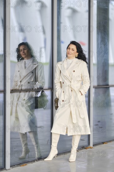 Dressed in a stylish white trench coat and matching boots, a woman stands confidently against a