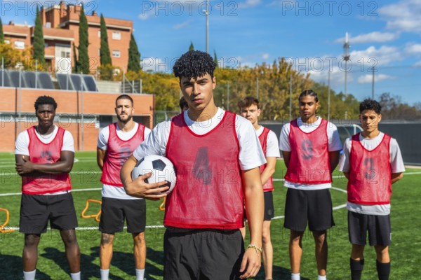 Multi ethnic group of young men in red bibs standing on a green football pitch, preparing for