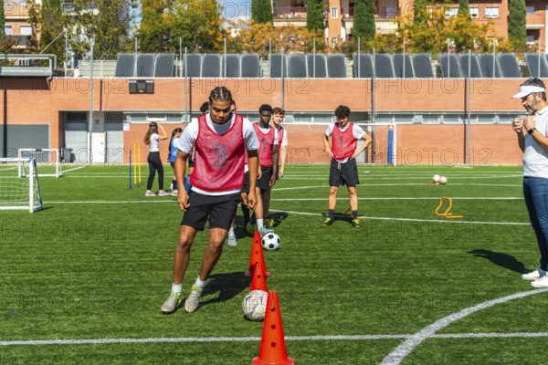 Young men in red vests are performing agility drills with cones and soccer balls on a green