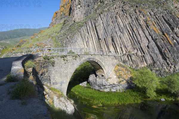 An old stone bridge stands in front of dramatic rock walls surrounded by green vegetation and a