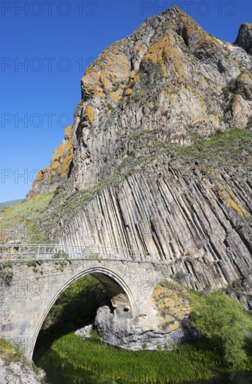 A high stone bridge stretches in front of an impressive rock formation under a bright blue sky,
