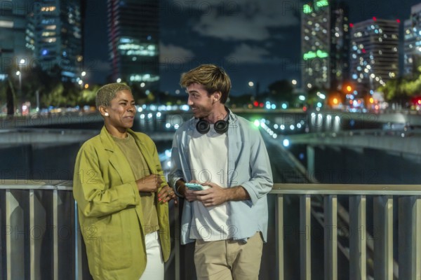Diverse young couple chatting and smiling on an urban bridge at night, city skyline and glowing