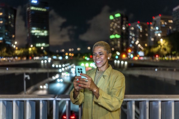 Woman standing on a city bridge at night, smiling while using a smartphone with blurred traffic and