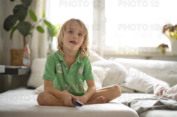 Child, boy, 7 years old, blonde, sitting on sofa, with remote control in hand, watching TV,