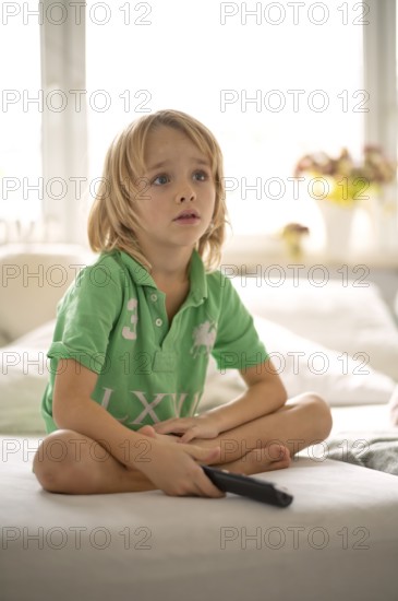 Child, boy, 7 years old, blonde, sitting on sofa, with remote control in hand, watching TV,