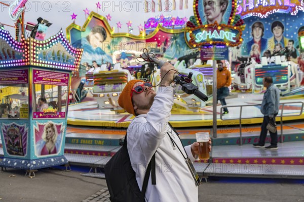 Influencer with live camera and beer in front of a fairground ride. Cannstatter Volksfest at the