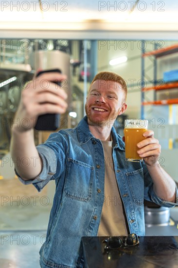 Smiling red haired man in a denim jacket takes a selfie with his smartphone while holding a glass