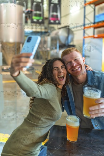 Happy young couple posing for a selfie with a smartphone, holding glasses of craft beer, and