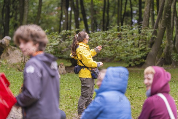 A person in a yellow jacket points to something in a wooded area. Surrounded by blurred figures,