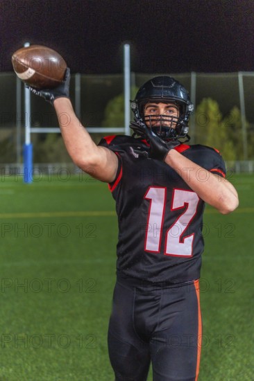 American football player in helmet and number 12 uniform holds the ball up on a green grass field