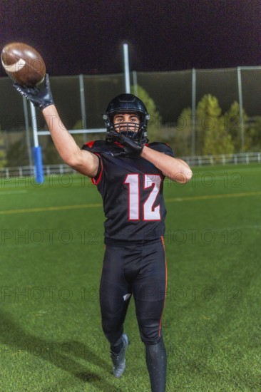 American football player wearing a black uniform with number 12 and helmet, extending arm to catch