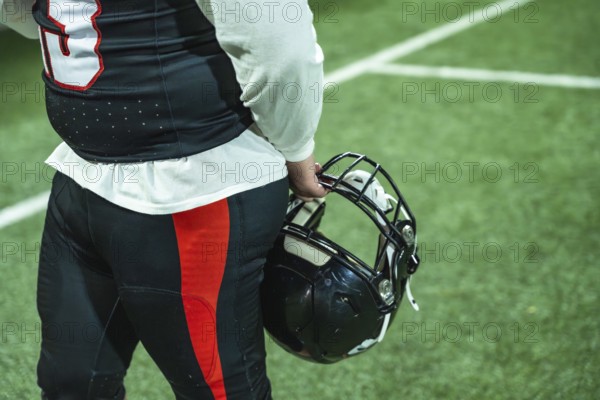 American football player standing on a green turf field, holding a black helmet and wearing a