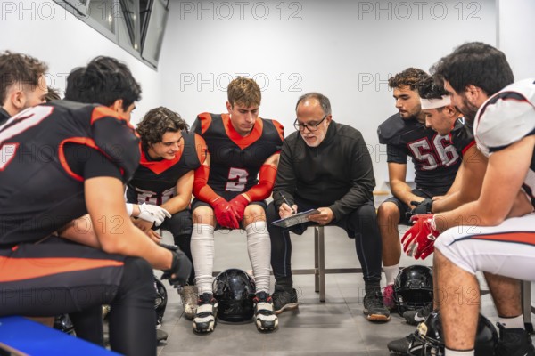 American football coach discussing strategy with his team of male players in the changing room,