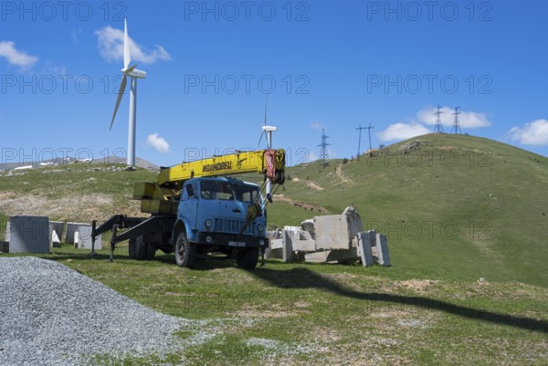 A blue crane lorry stands on a green construction site next to a wind turbine on a hill, Pushkin