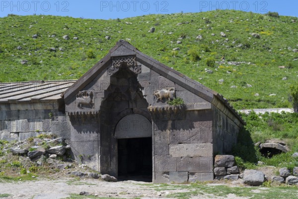 Old stone building with detailed decorations in front of a lush green hill on a sunny day,