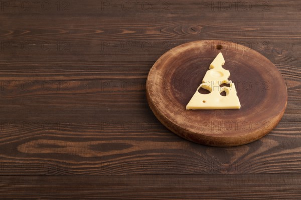 Piece of cheese with holes on cutting board on brown wooden background, side view, copy space,