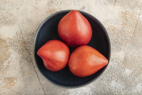 Red Heart shape tomatoes in blue bowl on brown concrete background. Top view, copy space, flat lay.