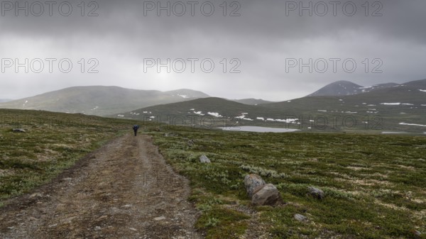 Hiker with rucksack and blue raincoat on a lonely plateau with snowfields, low clouds, Kongevegen