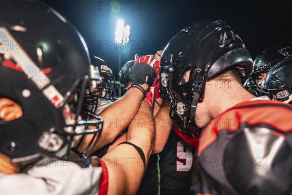 American football players are huddling on the field under stadium lights at night, putting their