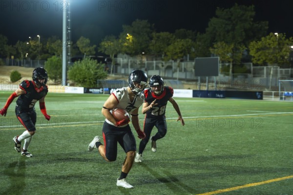 American football player vigorously running with the ball on a green field during a night game, two