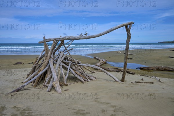 Treibholzskulptur Zelt am Strand unter blauem Himmel, ruhiges Meer im Hintergrund, Kaka Point,