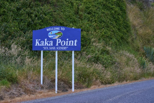 Willkommensschild von Kaka Point neben einer Straße mit grüner Vegetation und landschaftlicher