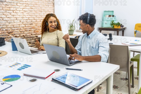Two smiling multiracial coworkers wearing headsets, collaborating and working on laptops at a