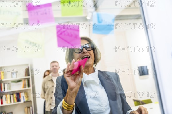 Mature businesswoman attaching colorful sticky notes on a clear glass board, collaborating on