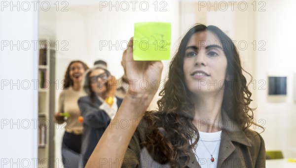 Young businesswoman sticking a green sticky note on a transparent glass wall during a team meeting,