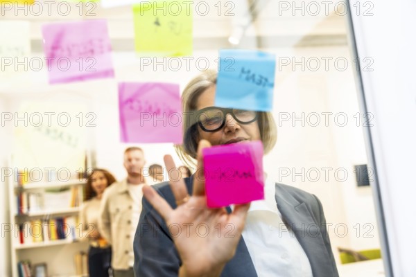 Mature businesswoman arranging colorful sticky notes on a glass board while leading a strategic