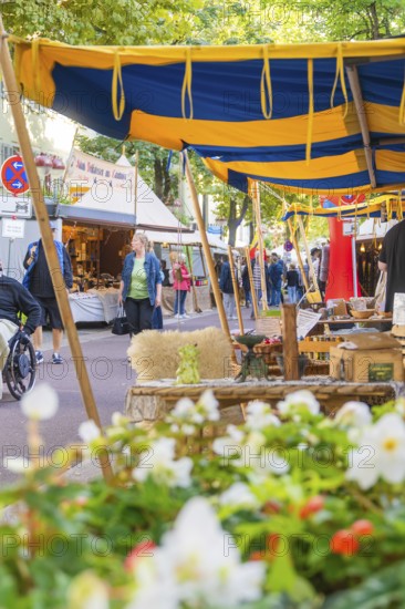 Lively market with blue and yellow tents, flowers in the foreground and many people shopping, Fire