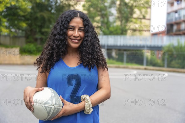 Young woman wearing a blue rugby uniform, confidently holding a rugby ball while smiling on an