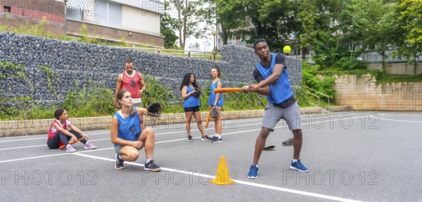 Multi ethnic baseball players practicing on an urban field, with a batter hitting the ball and a