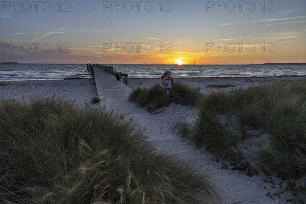 Sunset sky over the sea, wooden walkway and lifebuoy, framed by beach grass and calm water, Kobaek