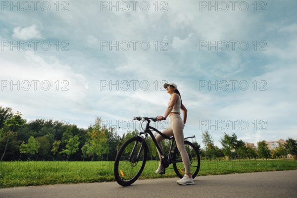A woman stands beside her bicycle in a lush green park. She wears comfortable athletic clothing and