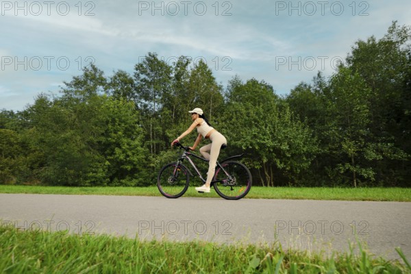 A woman rides her mountain bike on a smooth path in a lush green landscape. Tall trees line both