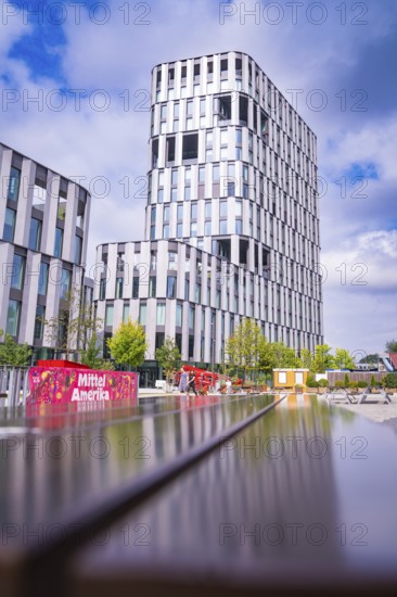 Futuristic high-rise building and park landscape under a blue sky with a colourful element, Munich,