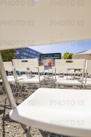 White chairs arranged in rows with a view of a screen and a building in the background, open air