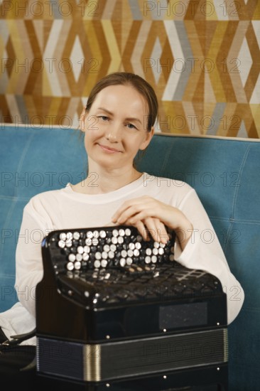 A middle-aged woman in a casual white top smiles, resting her hands on an accordion in a vibrant,
