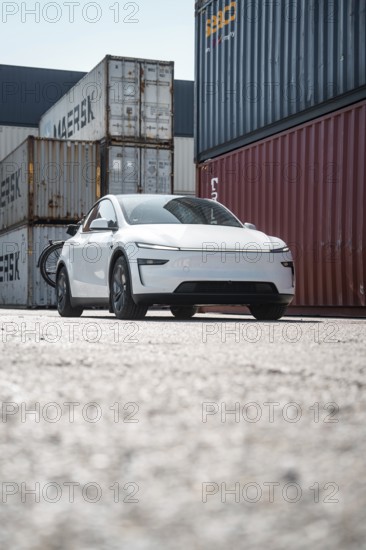 White car in a container port with different coloured freight containers, Tesla Model Y Juniper,